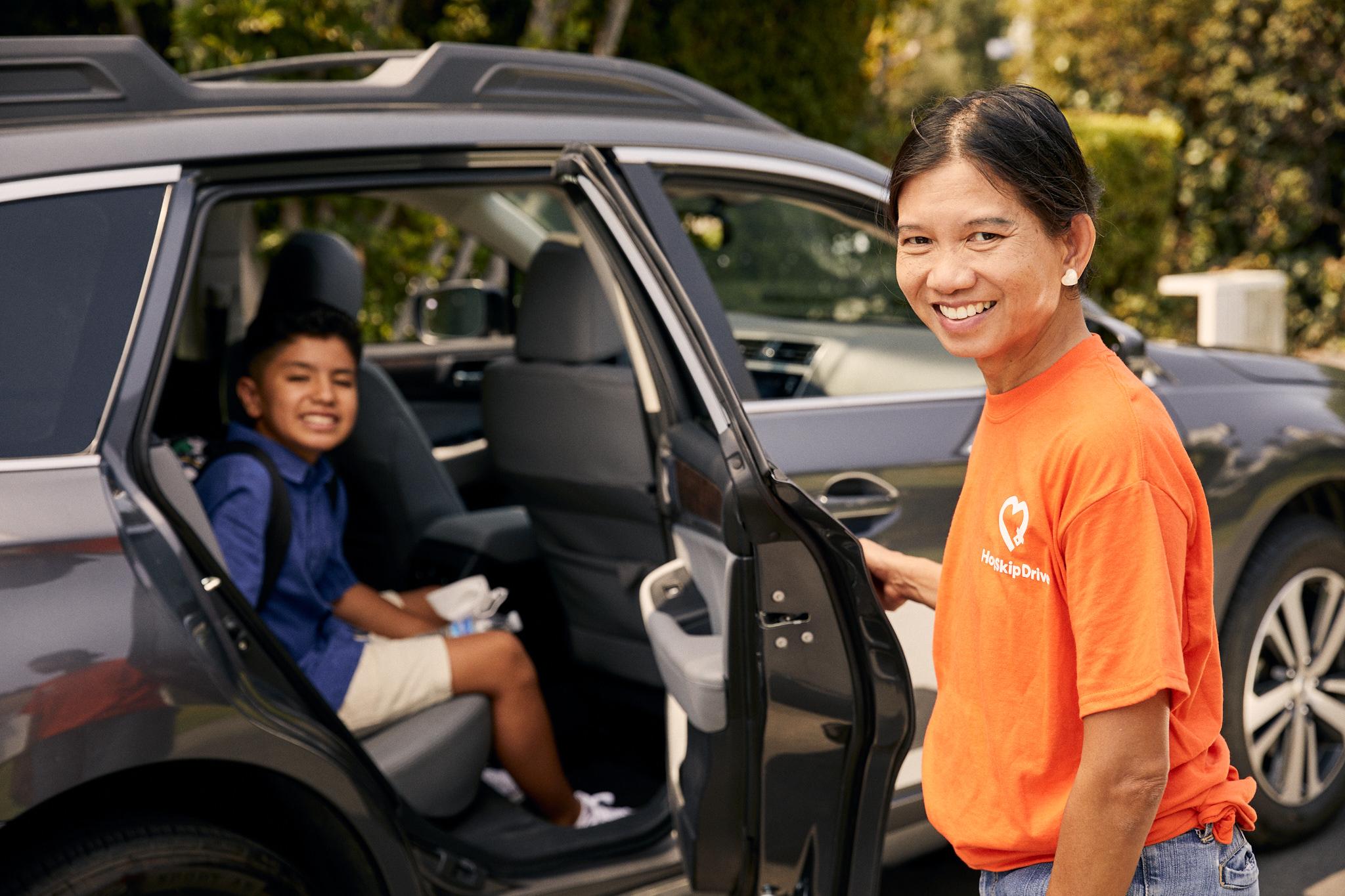 Driver in orange HopSkipDrive t-shirt standing by car door with young student passenger in school uniform seated inside
